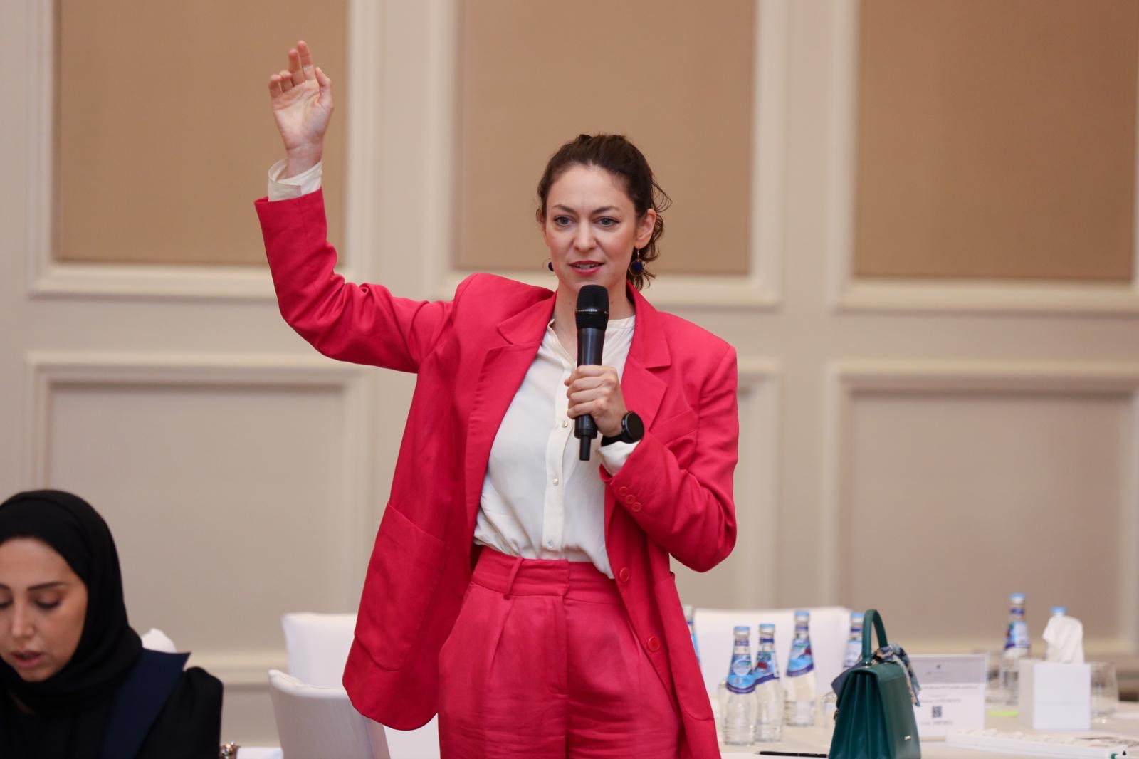 Lucy Antrobus in a red suit, arm raised, energising a workshop room in Doha