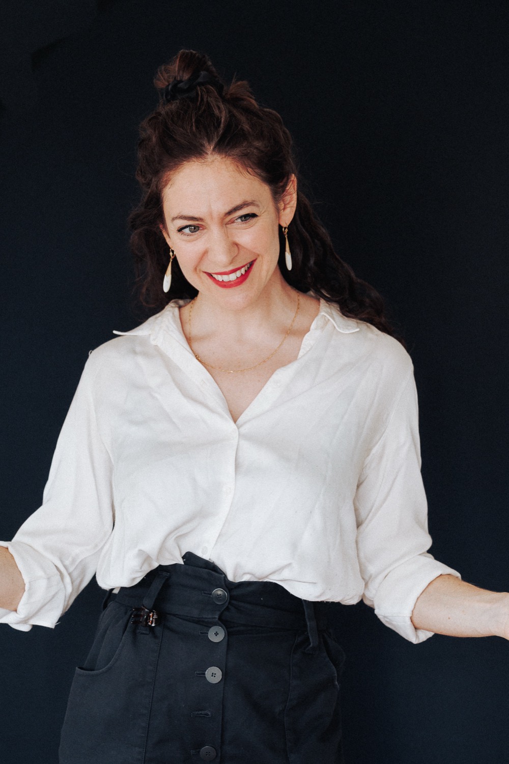 Lucy Antrobus smiling with arms open, in a white shirt against a dark background